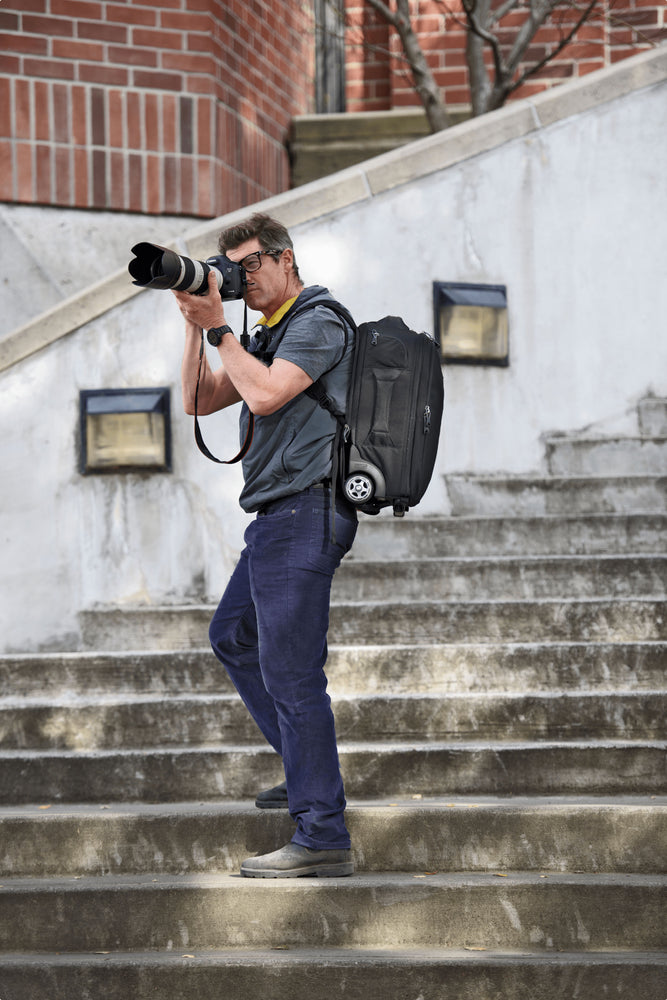 A man standing on the staircase wearing a roller backpack, holding the camera with long lens and getting ready to take a photo