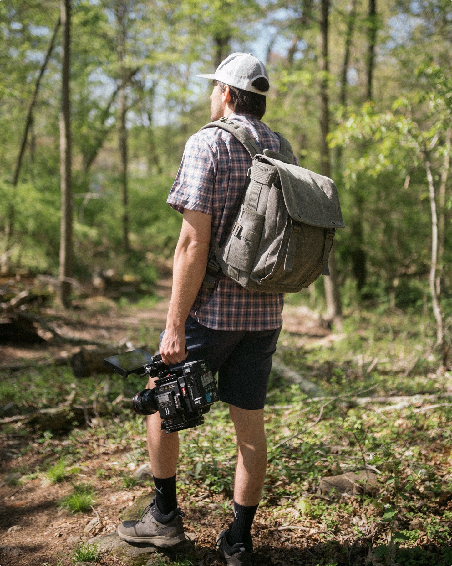 A man holding a filming equipment and wearing a backpack, gazing into the forest 