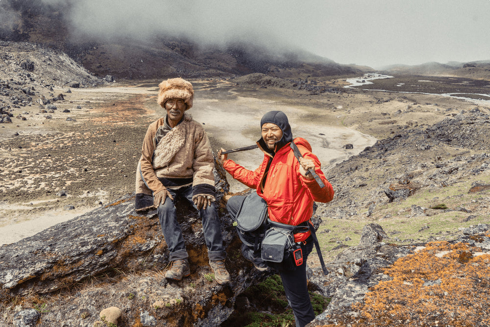 Two individuals sit on a mountainside, one in a fur hat and one in rain gear with camera equipment.