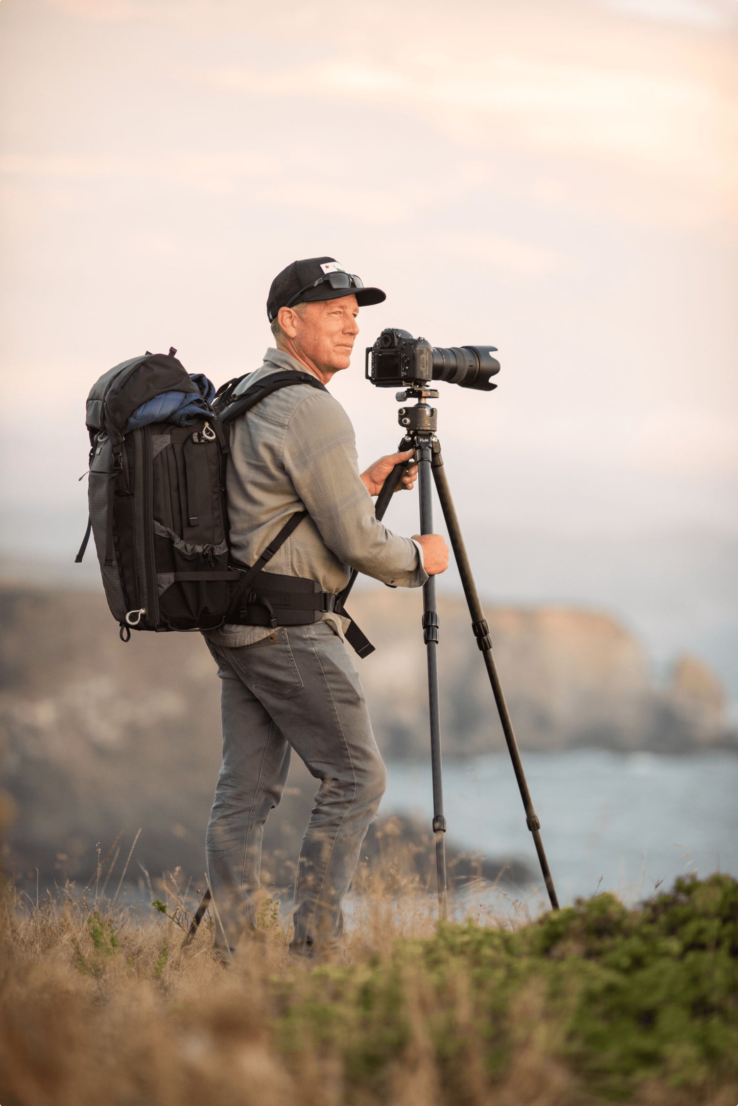 A photographer stands on a cliff wearing a backpack in front of a camera on a tripod.