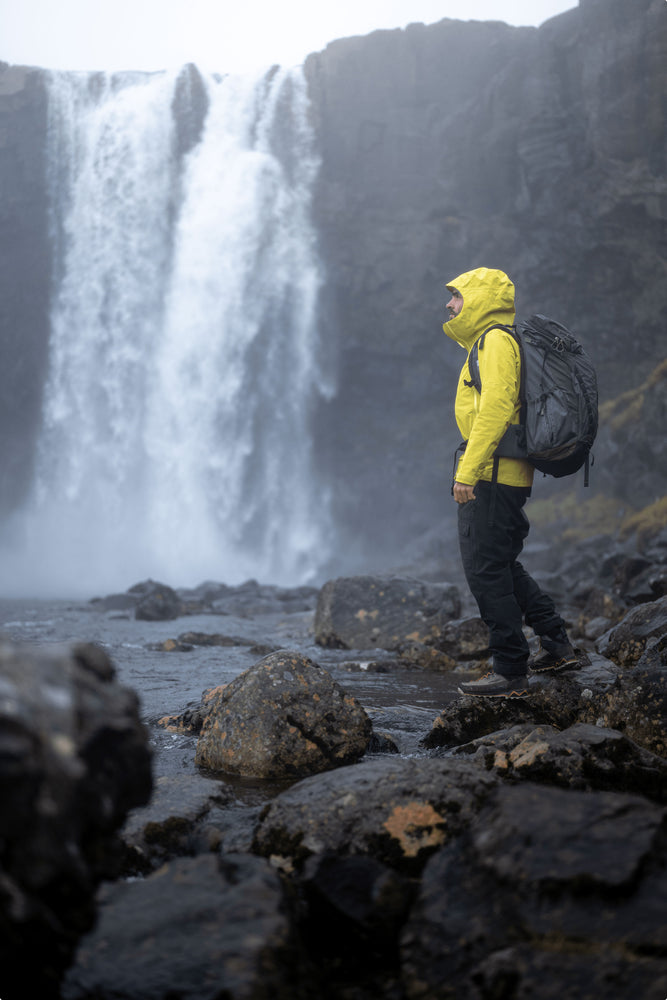 Highlands of Iceland scenery with a waterfall and a person balancing on nearby rocks wearing a raincoat and backpack